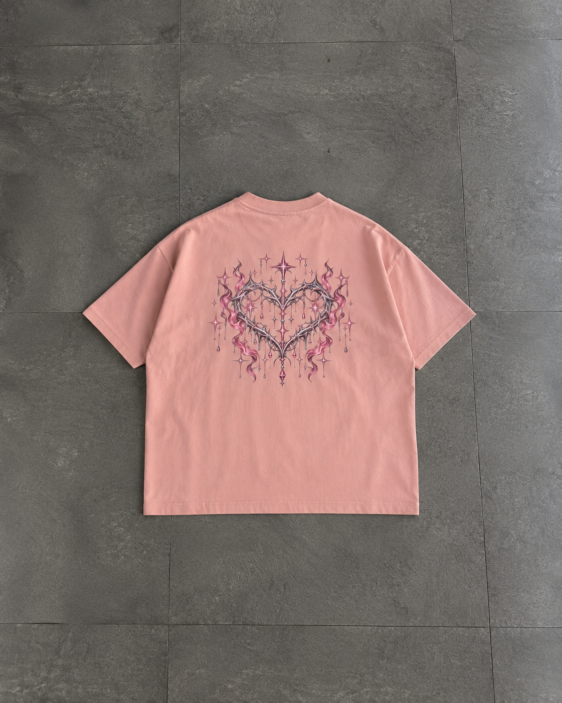 A light pink short-sleeve t-shirt laid flat on a dark gray tiled floor, featuring a large graphic print on the back with a heart and hanging star details.

Color: Peachy Back
