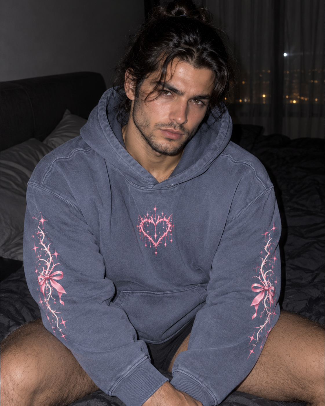 A man wearing a dark slate blue hoodie with pink embroidered floral and heart designs, sitting on a bed in a dimly lit bedroom.

Color: Dyed Blue Grey Front