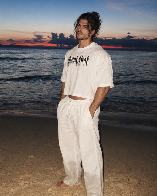 A man stands on a beach at sunset wearing a white cropped T-shirt with black gothic text reading 'Saint Laurent' and loose white drawstring pants.

Color: Oversized Fit