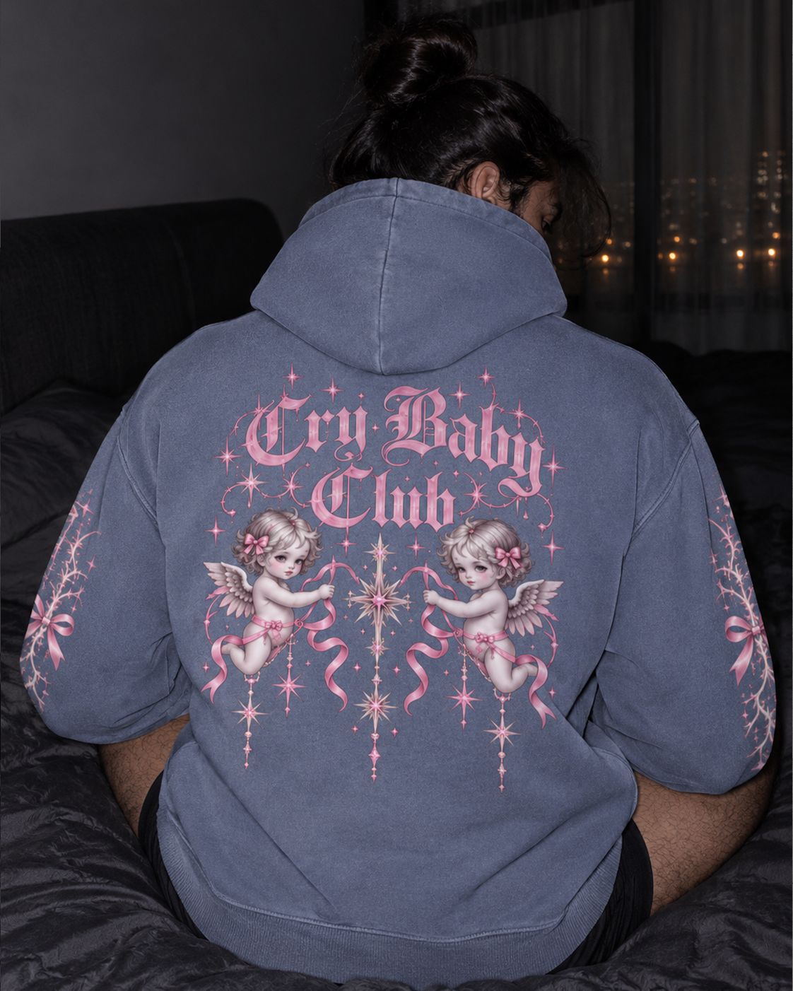 A person sitting on a bed wearing a faded slate blue hoodie from the back, featuring a large pink and white graphic with cherubs and the text 'Cry Baby Club'.

Color: Dyed Blue Grey Back