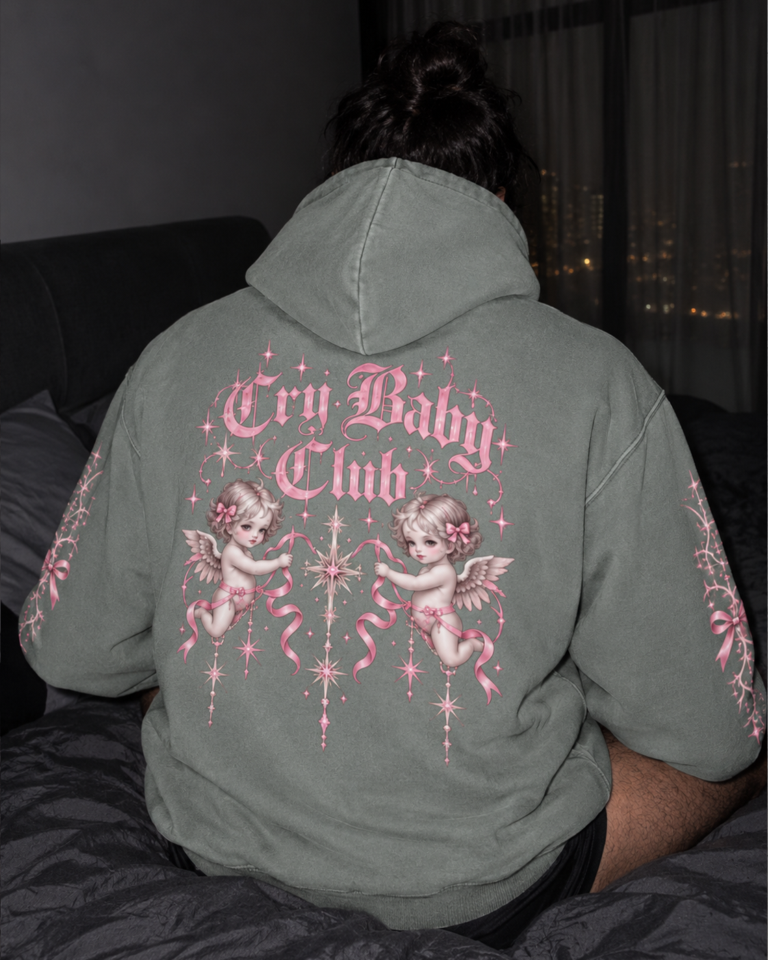 A person viewed from behind wearing an oversized muted sage green hoodie with a large pink and white 'Cry Baby Club' graphic featuring cherub illustrations on the back, sitting on a bed.

Color: Dyed Misty Grey Back