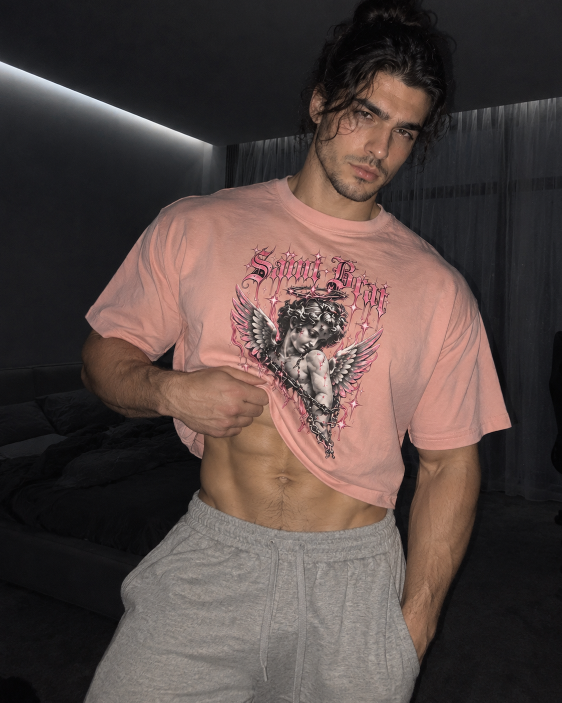 A man wearing a light pink graphic t-shirt with a cherub design and the text 'Saint Flicke', paired with gray sweatpants, in a dimly lit room.

Color: Peachy Front