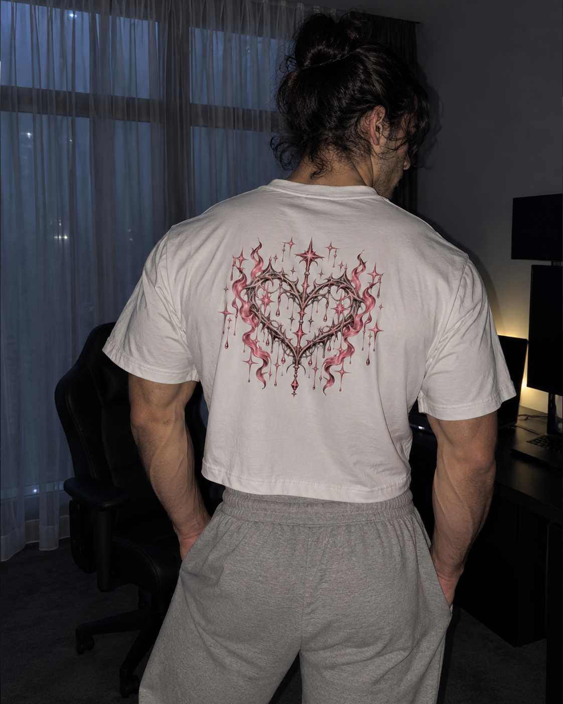 A man viewed from behind wearing a white t-shirt with a large red and black heart-shaped graphic on the back, paired with gray sweatpants in a dimly lit room.

Color: White Back