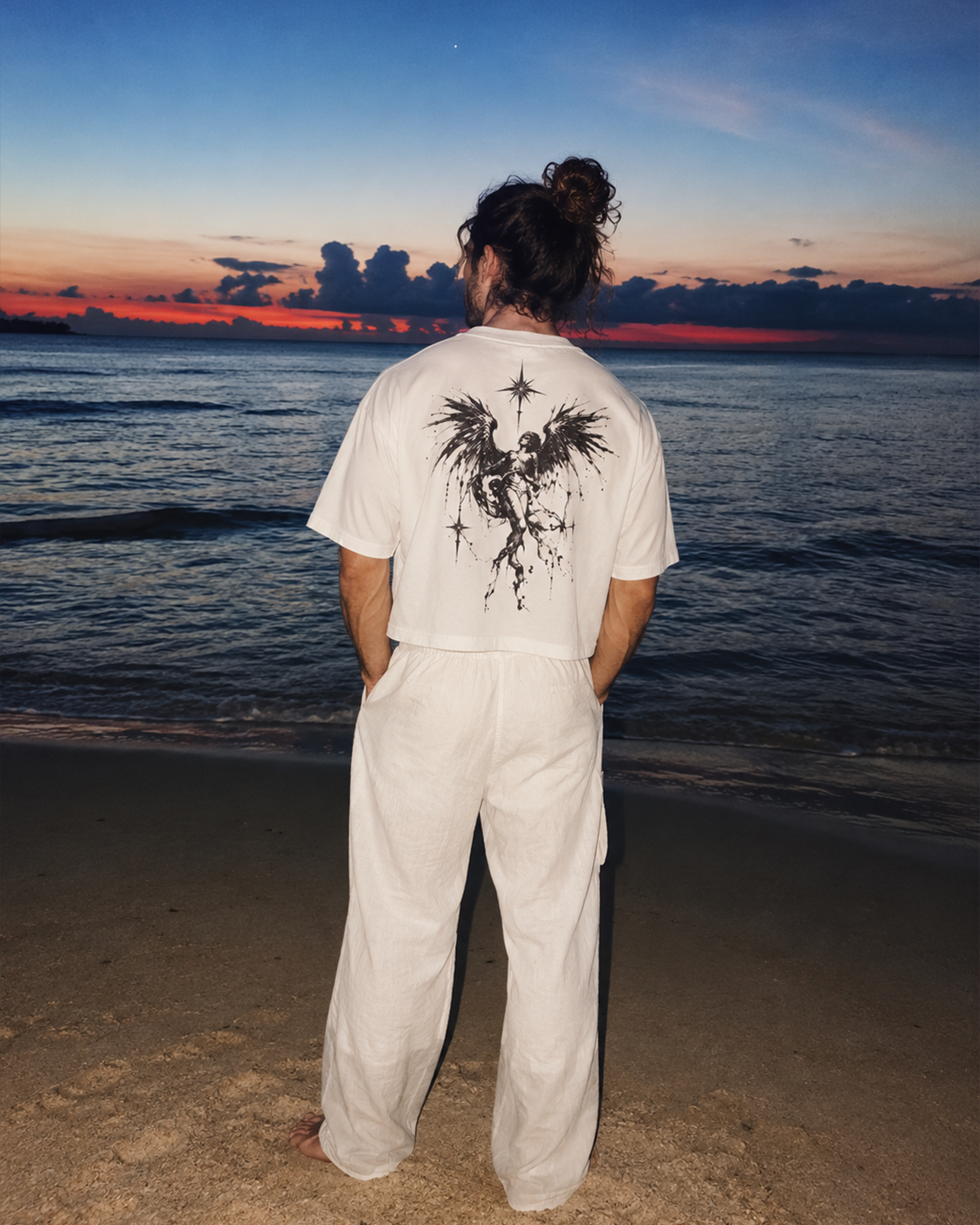 A person standing on a beach at sunset, viewed from behind, wearing a white short-sleeve T-shirt with a large black bird graphic on the back and matching white pants.

Color: Oversized Fit