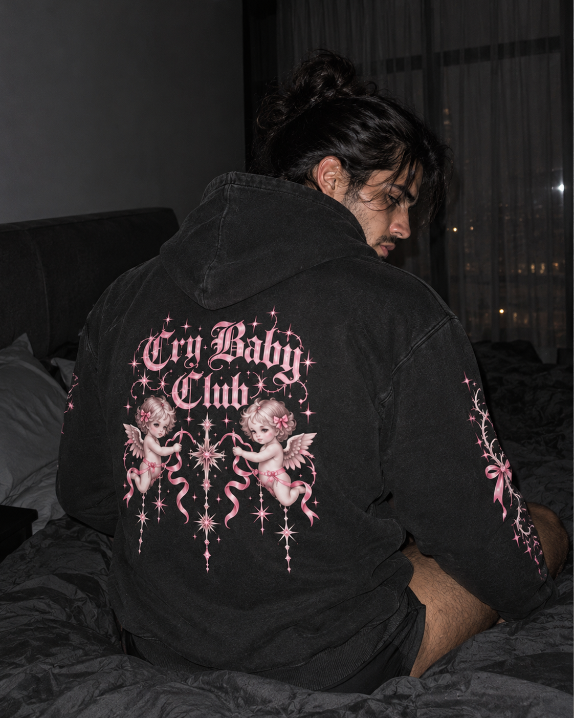 A person sitting on a bed wearing a black hoodie from the back, featuring a large pink and white 'Cry Baby Club' graphic with cherub illustrations on the back and matching graphics on the sleeves.

Color: Dyed Black Rock Back