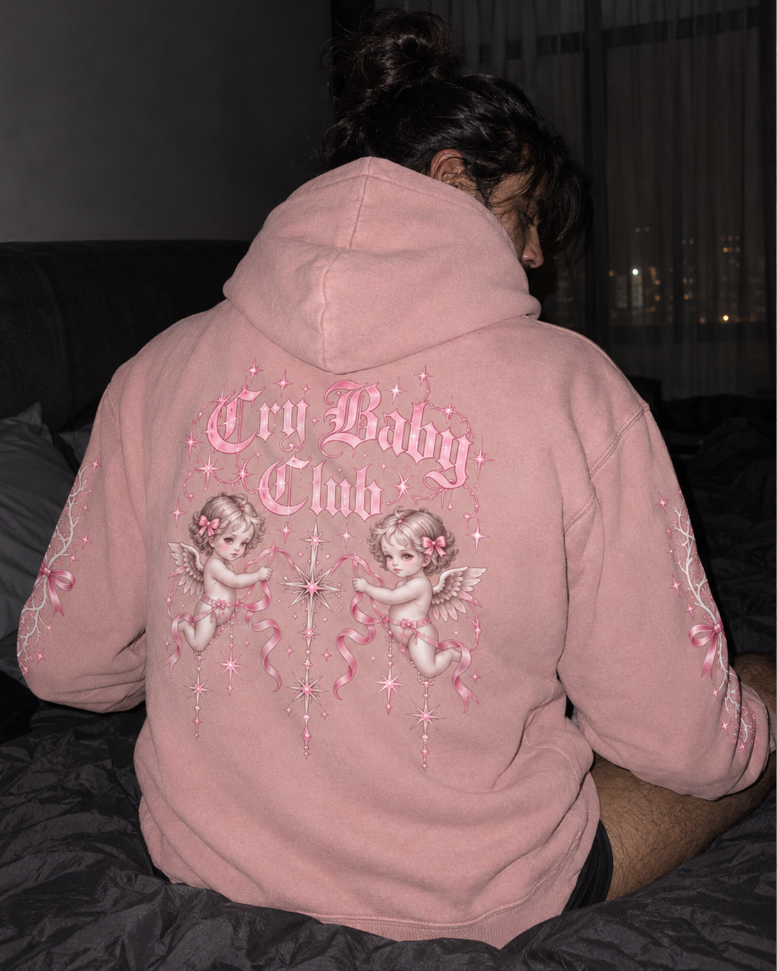A person sitting on a bed wearing a dusty pink hoodie from the back, featuring 'Cry Baby Club' text and cherub illustrations in pink and white embroidery.

Color: Dyed Blush Back