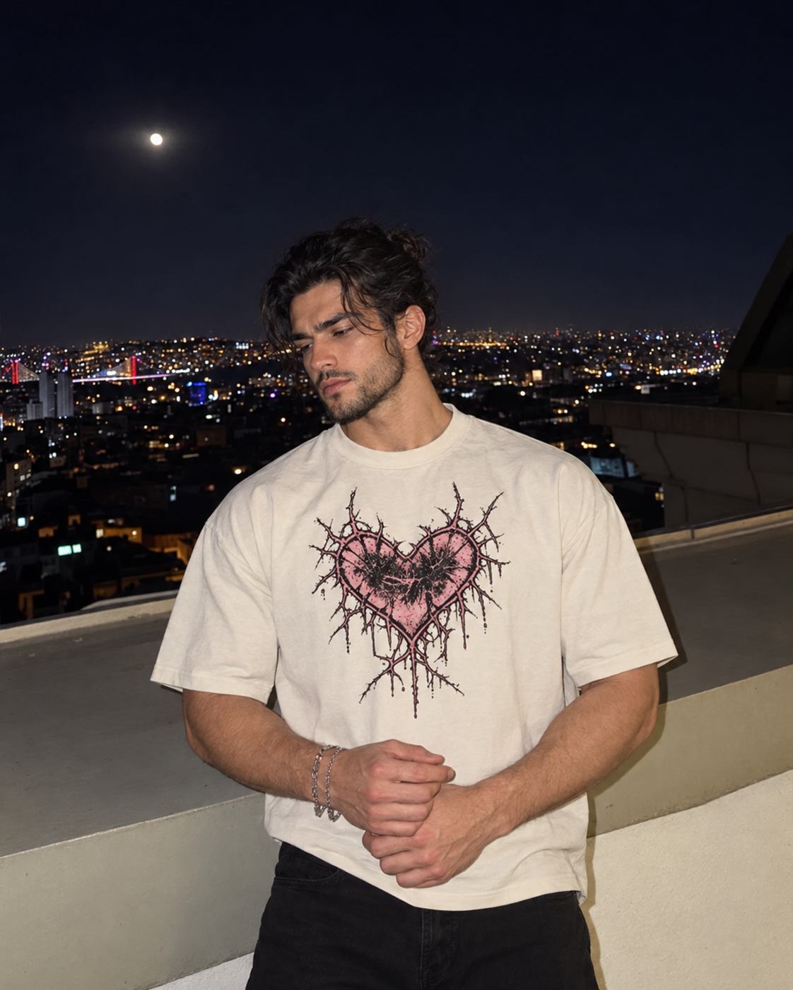 A man wearing a cream-colored t-shirt with a pink and black thorny heart graphic, standing on a rooftop at night with a city skyline and moon in the background.

Color: Vintage White Front