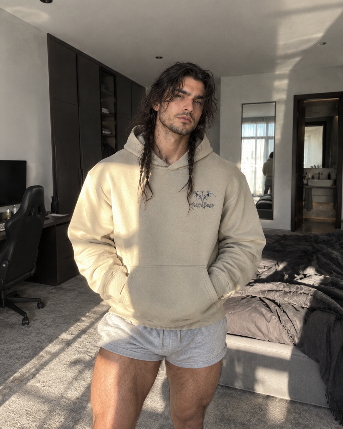 A man with long dark hair stands in a modern bedroom wearing a beige hoodie with a small embroidered graphic on the left chest and light gray shorts.
Color: Stone Front 
