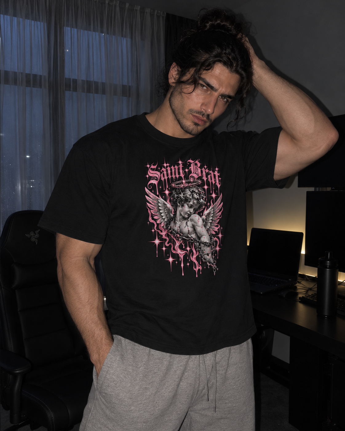 A man wearing a black Saint Patrick gothic graphic t-shirt with a pink and white cherub design, paired with gray sweatpants, standing in a dimly lit room.

Color: Black Front