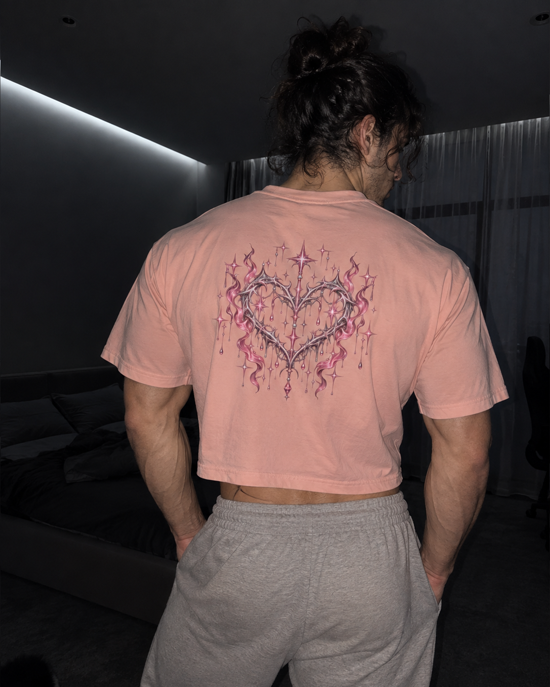 A muscular man viewed from behind wearing a light pink cropped t-shirt with a large pink and purple heart-shaped graphic on the back, paired with gray sweatpants in a dark bedroom setting.

Color: Peachy Back