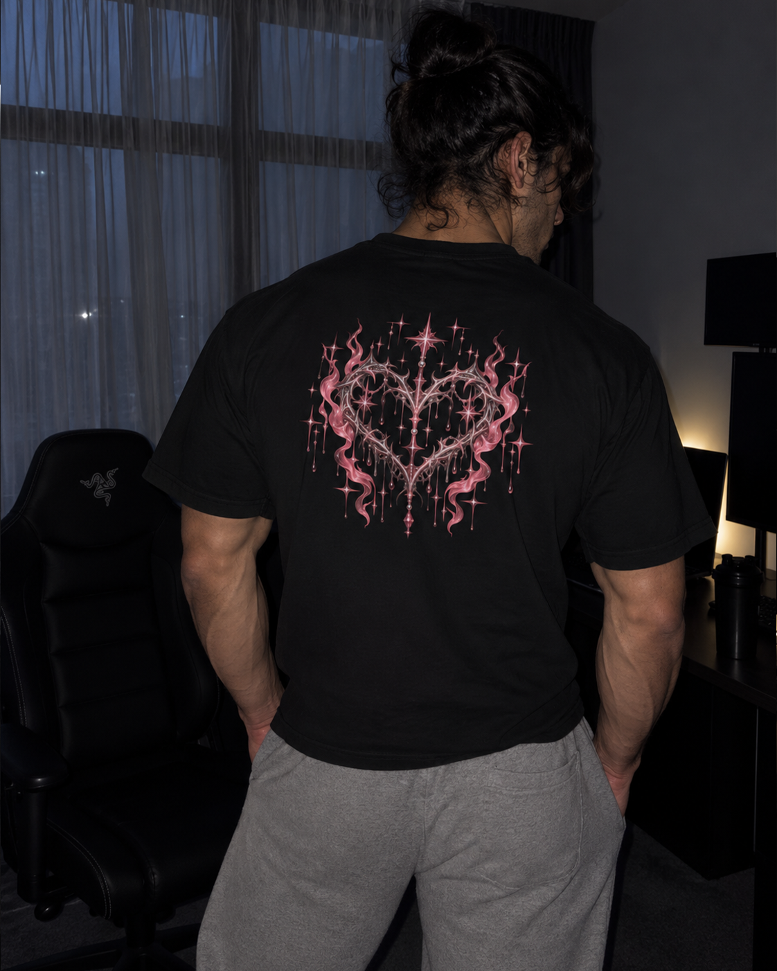 A man viewed from behind wearing a black t-shirt with a large pink and red heart-shaped graphic print on the back, paired with gray sweatpants in a dimly lit room.

Color: Black Back 