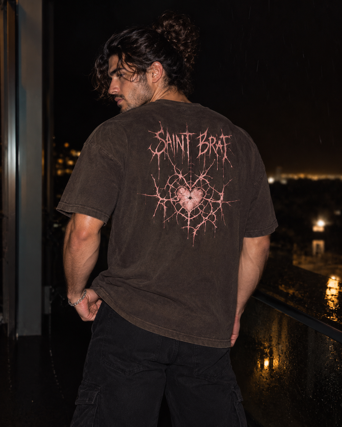 A man wearing a dark brown washed t-shirt with a pink and white 'Saint Brat' graphic on the back, standing on a balcony at night.

Color: Vintage Black Back