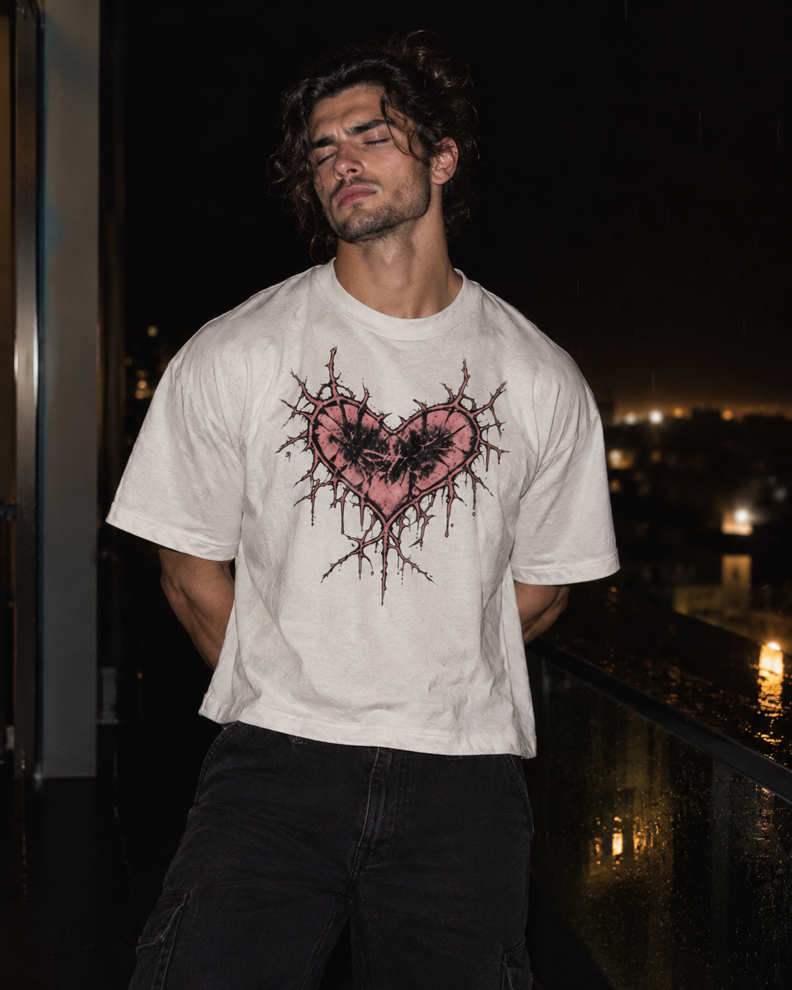 A man wearing a white graphic t-shirt with a pink and black heart design, standing on a balcony at night.

Color: White Front
