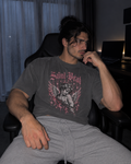 A man sitting in a black office chair wearing a dark gray graphic t-shirt with a pink and white 'Saint Brat' cherub design, paired with light gray sweatpants in a dimly lit room.

Color: Pepper Front