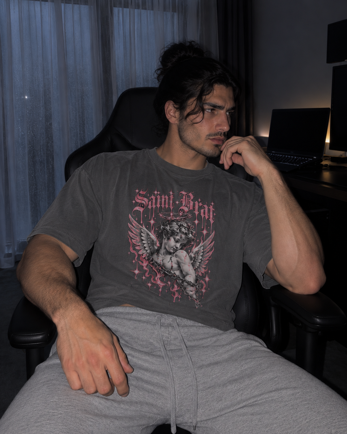 A man sitting in a black office chair wearing a dark gray graphic t-shirt with a pink and white 'Saint Brat' cherub design, paired with light gray sweatpants in a dimly lit room.

Color: Pepper Front