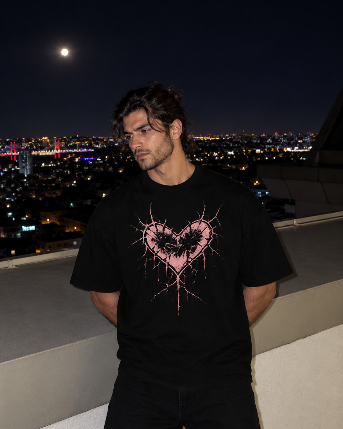 A man wearing a black t-shirt with a pink heart graphic, standing on a rooftop at night with a city skyline and full moon in the background.

Color: Black Front 