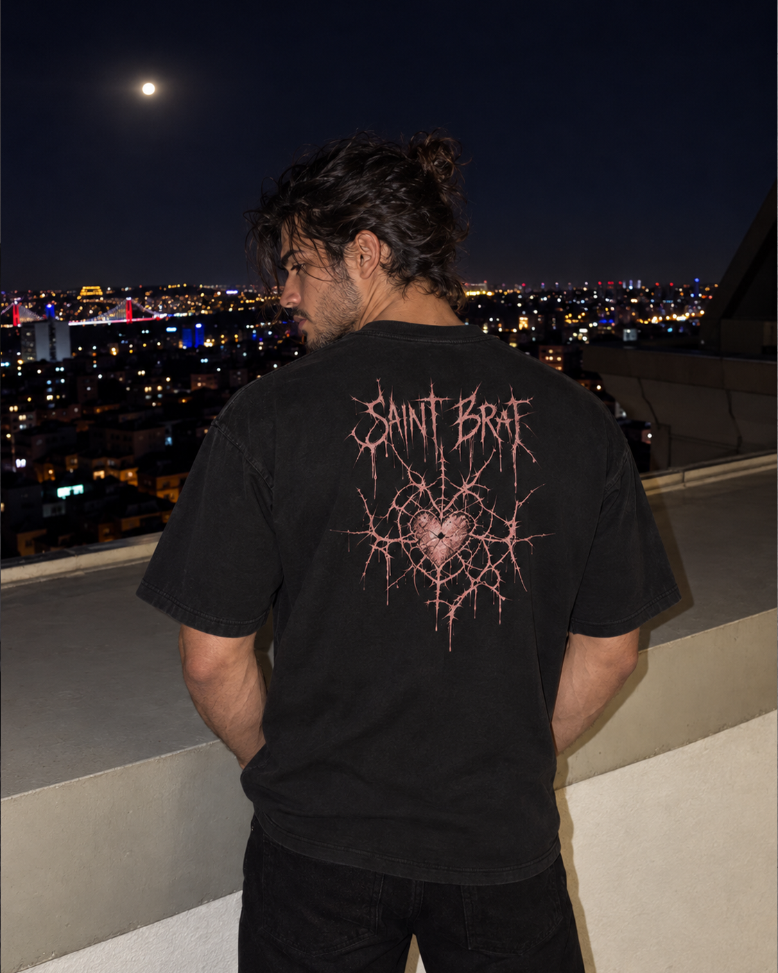 A man stands on a rooftop at night wearing a black t-shirt with a pink gothic-style graphic on the back that reads 'SAINT BART'. The background shows a city skyline illuminated by lights under a full moon.

Color: Black Back