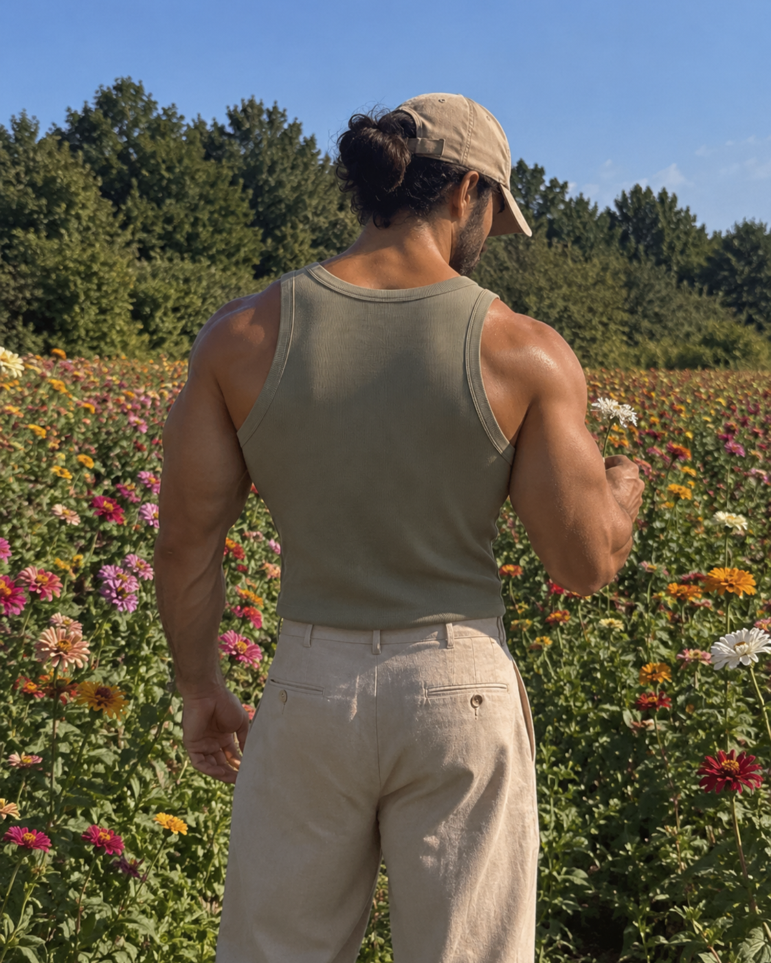 A muscular man viewed from behind wearing an olive green ribbed tank top, beige pants, and a tan baseball cap, standing in a vibrant flower field with trees and blue sky in the background.

Color: Eucalyptus Back