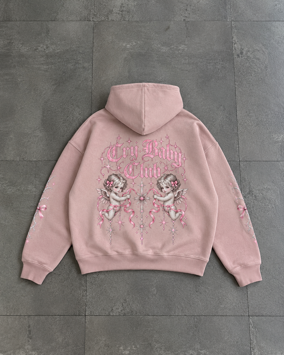 A light pink hoodie laid flat on a dark gray tiled floor, showing the back design with 'Cry Baby Club' text, cherub illustrations, and pink ribbon graphics.

Color: Dyed Blush Back