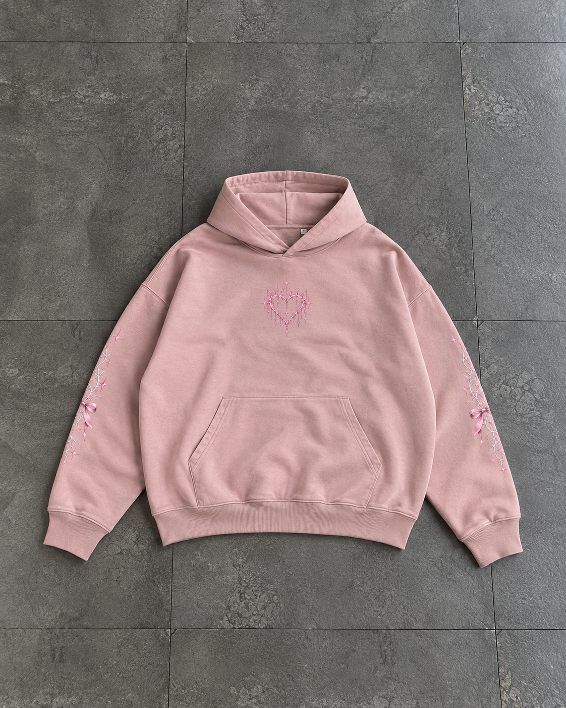 A light pink pullover hoodie with a small pink heart design on the chest and matching pink floral embroidery on both sleeves, laid flat on a dark gray tiled surface.

Color: Dyed Blush Front