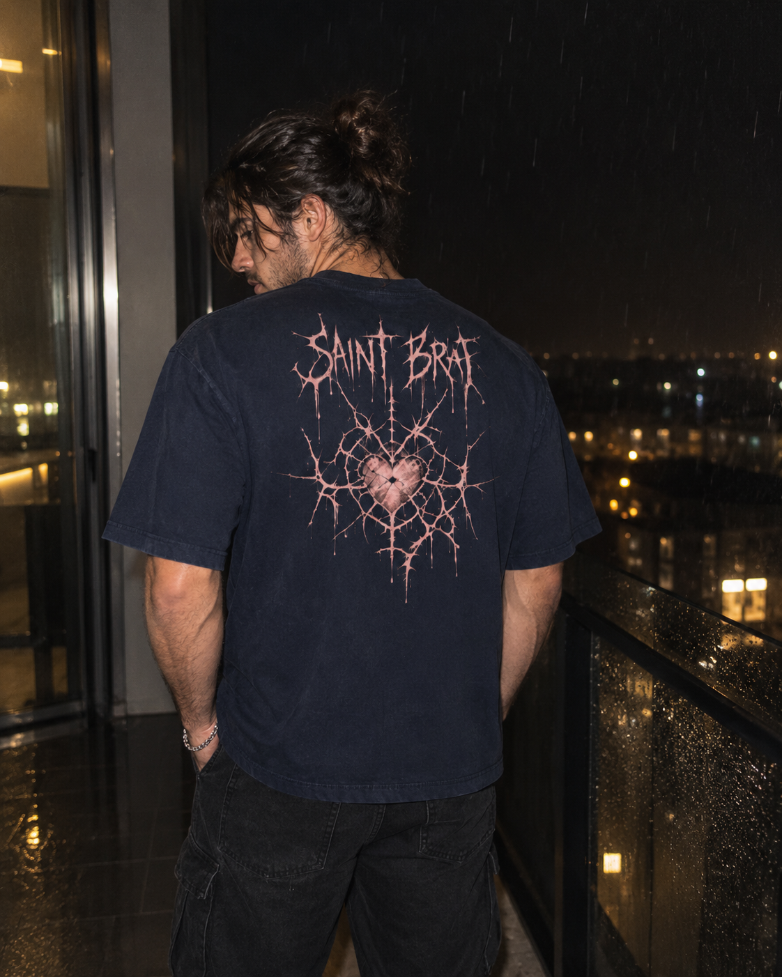 A man stands on a balcony at night wearing a dark navy blue t-shirt with a large pink graphic on the back. The graphic features the text 'SAINT BRAT' in a distressed, metallic font above a cracked pattern surrounding a heart-shaped graphic.

Color: Navy Blazer Back