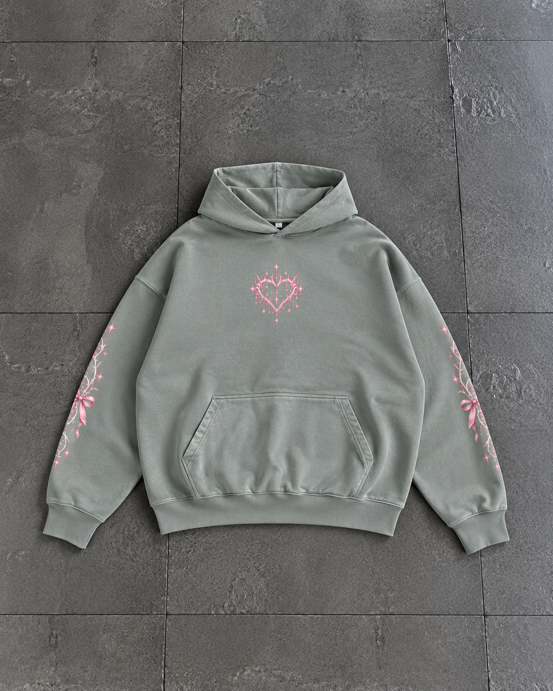 A sage green pullover hoodie with a pink heart graphic on the chest and pink floral patterns on both sleeves, laid flat on a dark gray tiled surface.

Color: Dyed Misty Grey Front