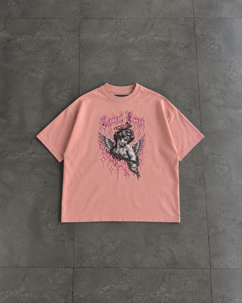A light pink short-sleeve t-shirt with a black and pink graphic of a winged cherub, laid flat on a dark gray tiled surface.

Color: Peachy Front