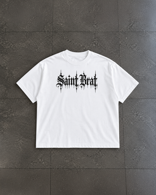 A white short-sleeve crewneck t-shirt with a black gothic-style 'Saint Beast' graphic printed across the chest, laid flat on a dark gray tiled surface.