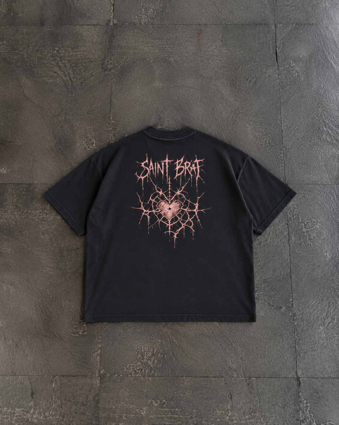 A black short-sleeve t-shirt laid flat on a dark gray textured stone surface, featuring a pink and white graphic on the back with the text 'SAINT BFAI' and a cracked heart design.

Color: Black Back