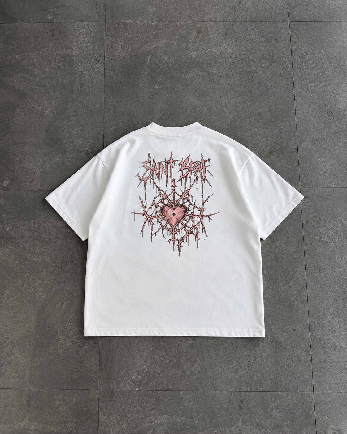 A white short-sleeve t-shirt laid flat on a dark gray tiled floor, showing a back graphic with the text 'Saint Sinner' in a jagged, thorny style above a pink heart.

Color: White Back