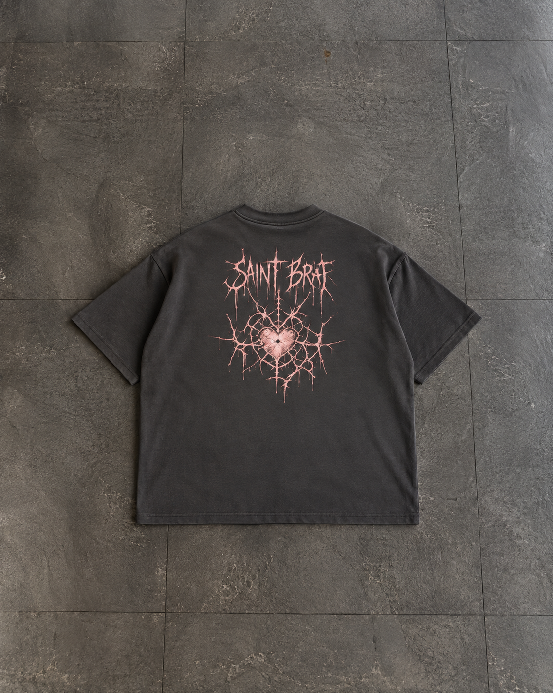 A dark charcoal gray t-shirt laid flat on a dark gray tiled floor, featuring a pink and white graphic on the back with the text 'SAINT BATA' and a cracked heart design.

Color: Vintage Black Back