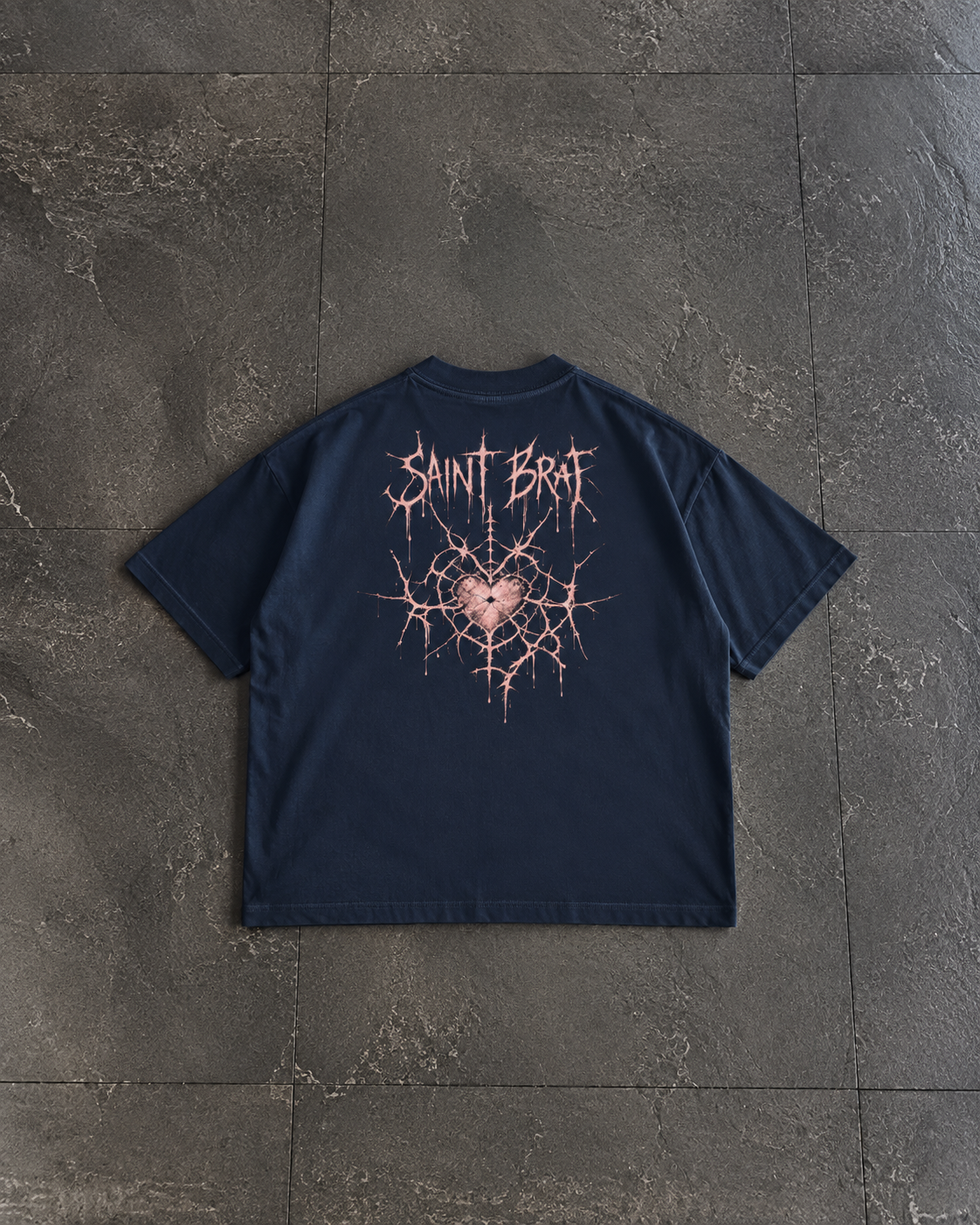 A navy blue short-sleeve t-shirt laid flat on a dark gray tiled surface, featuring a distressed pink graphic on the back with the text 'SAINT BFAI' above a cracked heart design.

Color: Navy Blazer Back