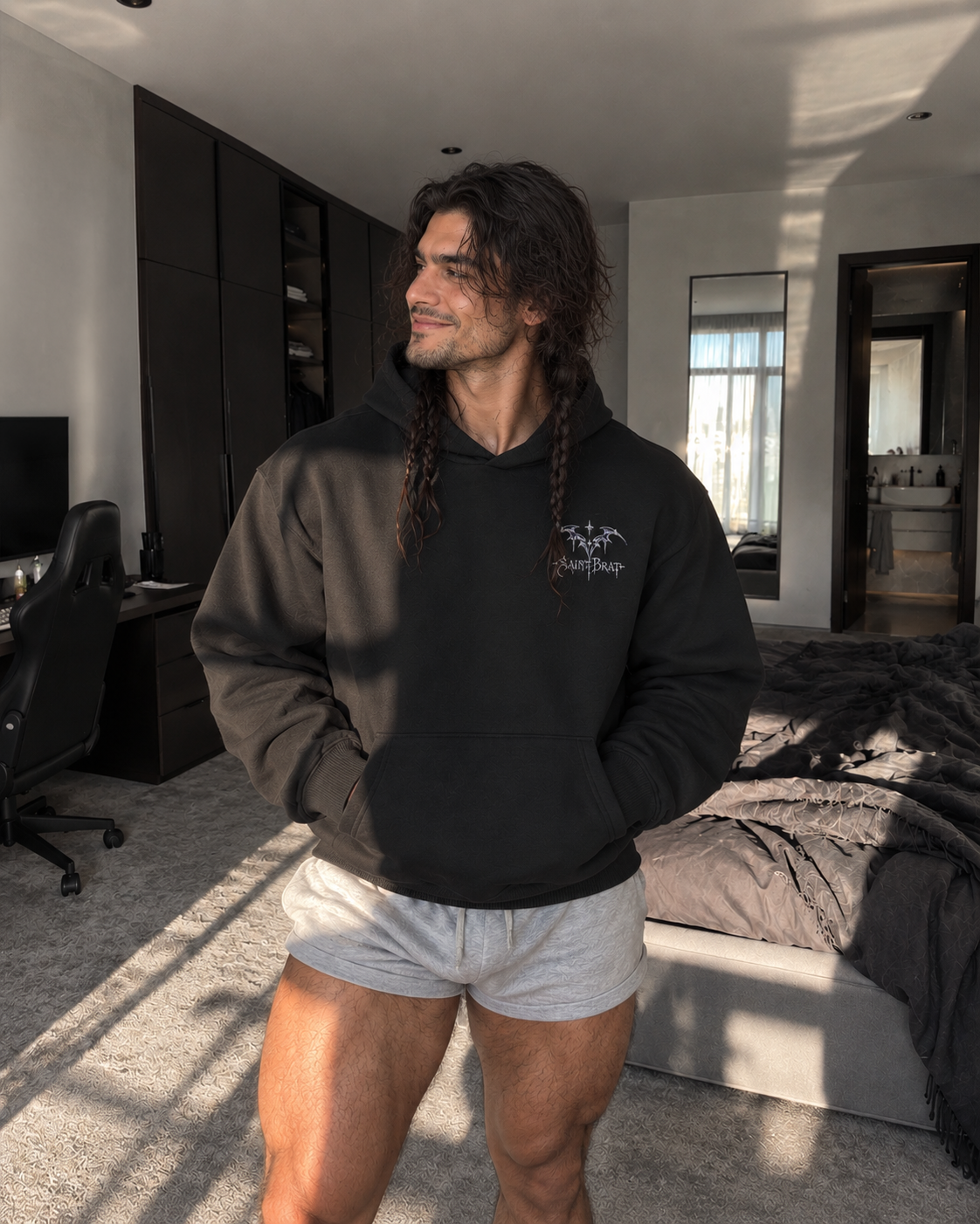 A man with long dark hair stands in a modern bedroom wearing a black pullover hoodie with a small white chest graphic and light gray shorts.

Color: Black Front 